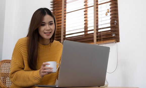 Young woman relaxing and drinking cup of hot coffee or tea using laptop computer on chair.woman checking social apps and working.Communication and technology concept.