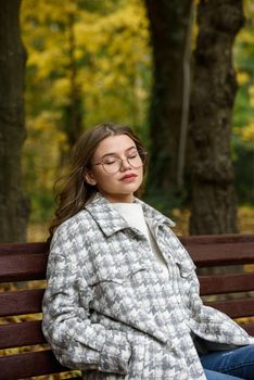 European elegant young woman in a stylish trench coat in a white sweater and blue jeans resting on the bench. warm autumn day. brown wooden bench