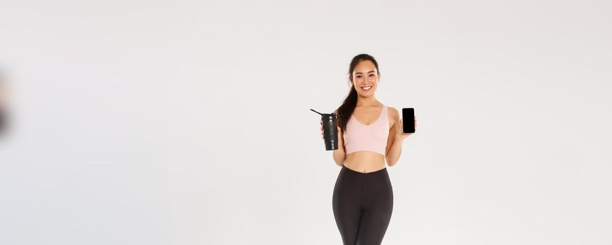 Full length of smiling good-looking asian fitness girl, sportswoman in activewear showing smartphone screen and water bottle, control drinking liquid with mobile application during training session.