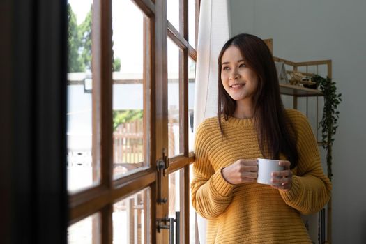 Happy young woman at home standing near window relaxing in her living room and drinking a cup of coffee..