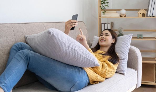 Young asian woman is using smartphone lie down on the sofa. Relax, happy, technology concept.