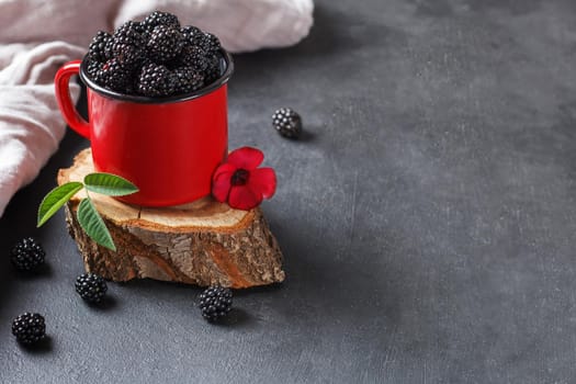 Fresh blackberry in a mug on a wooden stand on a black background with a flower. The concept of natural food. copy space