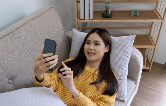 Young asian woman is using smartphone lie down on the sofa. Relax, happy, technology concept.