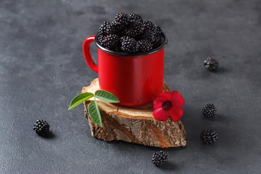 Fresh blackberry in a mug on a wooden stand on a black background with a flower. The concept of natural food.