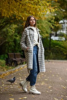 European elegant young woman in a stylish trench coat in a white sweater, blue jeans and white boots walking in a park. warm autumn day.