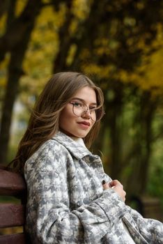 Close-up portrait of young woman in a stylish trench coat and in a white sweater resting on the bench. warm autumn day. warm autumn day. brown wooden bench