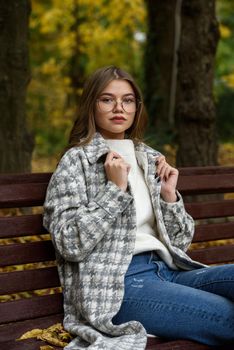 Close-up portrait of young woman in a stylish trench coat and in a white sweater resting on the bench. warm autumn day. warm autumn day. brown wooden bench