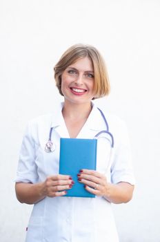 Young beautiful caucasian woman doctor with a phonendoscope on her neck and with a notepad smiling on a light background. High quality photo