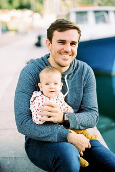 Happy dad with a baby on his knees sits on the pier. High quality photo