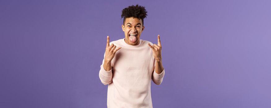 Portrait of enthusiastic, excited funny young man having fun, enjoying awesome rock concert, show heavy metal sign, stick tongue and rejoicing hear favorite band on stage, purple background.