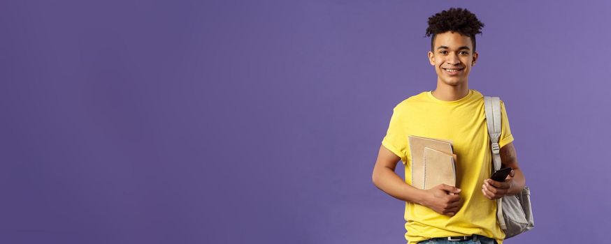 Back to school, university concept. Portrait of young cheerful male student with dreads, hipster going to his campus, carry backpack and study material, notebooks, purple background.