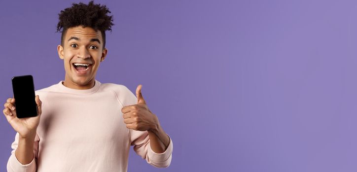 Happy young man, hispanic guy with dreads recommend app, online delivery service or food order, show thumbs-up, smiling excited, holding mobile phone, show smartphone screen.