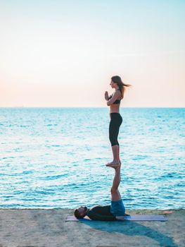 Young beautiful couple practicing acro yoga on the sea beach near water. Man and woman doing everyday practice outdoor on nature background. Healthy lifestyle concept.