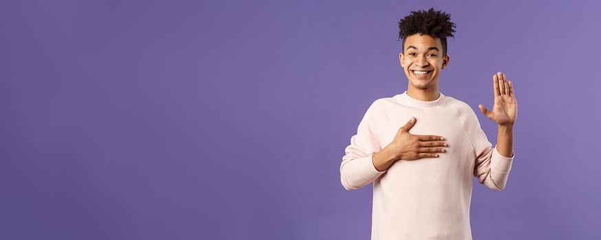 Portrait of young happy, joyful hispanic man making honest statement, promise to tell only truth, hold one hand on heart and another raised, smiling sincere, standing purple background.