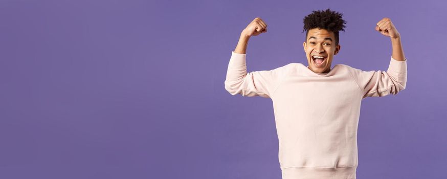 Portrait of young happy man got scholarship, applied to cool university, raise hands up flex biceps like champion, triumphing from great news, achieve goal and rejoicing, purple background.