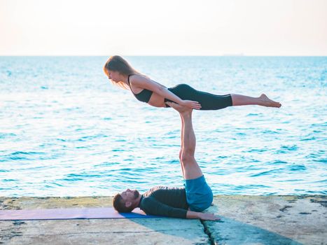 Young beautiful couple practicing acro yoga on the sea beach near water. Man and woman doing everyday practice outdoor on nature background. Healthy lifestyle concept.