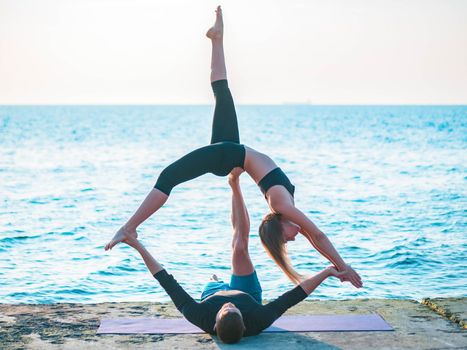 Young beautiful couple practicing acro yoga on the sea beach near water. Man and woman doing everyday practice outdoor on nature background. Healthy lifestyle concept.