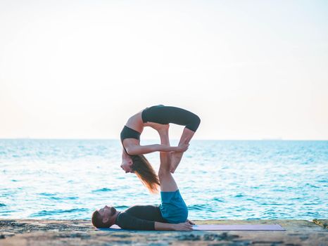 Young beautiful couple practicing acro yoga on the sea beach near water. Man and woman doing everyday practice outdoor on nature background. Healthy lifestyle concept.