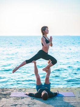 Young beautiful couple practicing acro yoga on the sea beach near water. Man and woman doing everyday practice outdoor on nature background. Healthy lifestyle concept.