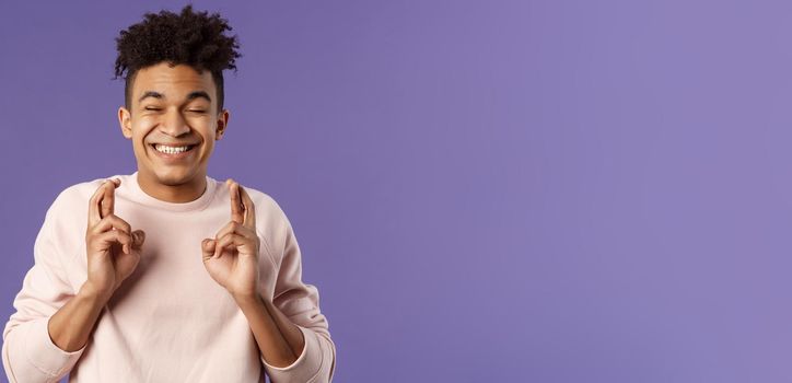 Close-up portrait of happy young hopeful guy anticipating something good happen, cross fingers good luck, close eyes and smiling awaiting miracle, praying for dream come true, purple background.