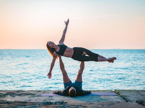 Young beautiful couple practicing acro yoga on the sea beach near water. Man and woman doing everyday practice outdoor on nature background. Healthy lifestyle concept.