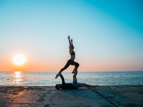 Young beautiful couple practicing acro yoga on the sea beach near water. Man and woman doing everyday practice outdoor on nature background. Healthy lifestyle concept.