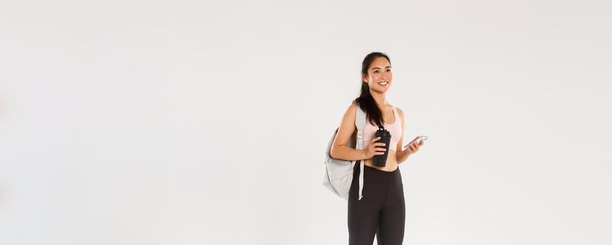 Full length of smiling healthy, slim asian girl going fitness training, female athelte carry backpack with workout equipment and water bottle, using mobile phone sports application, white background.