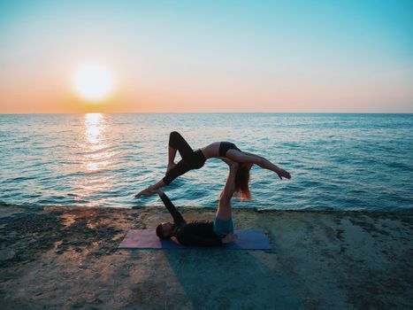 Young beautiful couple practicing acro yoga on the sea beach near water. Man and woman doing everyday practice outdoor on nature background. Healthy lifestyle concept.