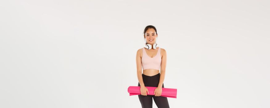 Full length of smiling slim asian girl with rubber mat for fitness exercises, headphones, looking at camera while waiting for training coach starting workout session in gym, white background.