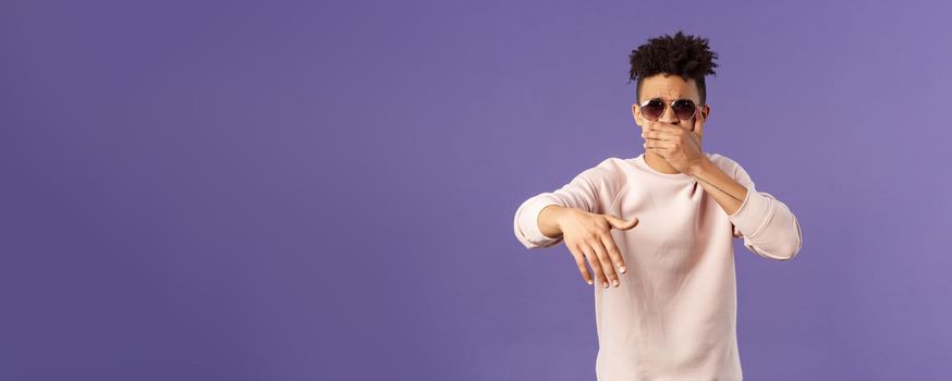 Waist-up portrait of cool and sassy, young carefree guy with dreads and sunglasses, cover mouth to beatbox, waving hand in rhythm music, singing rap or attend hip-hop party, purple background.