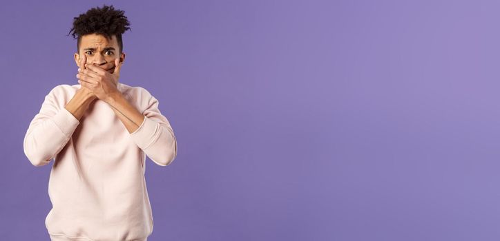 Shocked worried, embarrassed young man said something he shouldnt have, shut his mouth with hands and look guilty or anxious at camera, feel sorry for being rude, purple background.