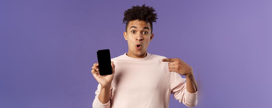 Close-up portrait of excited, cheerful young hispanic male geek talking about his device, new gadget or application, holding mobile phone, pointing at smartphone with amused face.