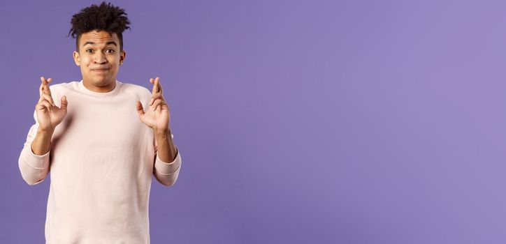 Portrait of young hopeful handsome man with dreads ancitipating something good happen, cross fingers good luck, smiling with joy and optimism, having faith, praying over purple background.