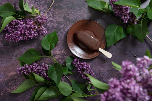 raspberry ice cream in chocolate on a wooden stick on a dark burgundy background with lilac flowers, a sweet dessert on a hot spring day, flat lay food. High quality photo