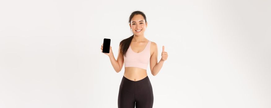 Sport, technology and active lifestyle concept. Satisfied smiling asian fitness girl, attractive sportswoman showing thumbs-up and smartphone screen, recommend mobile workout tracker app.