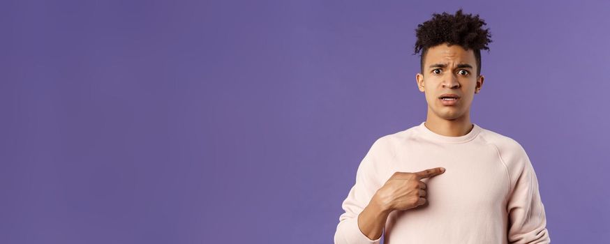 Close-up portrait of embarrassed and worried young man being named or called to principal office, pointing at himself and asking why with disbelief and anxiety, standing purple background.