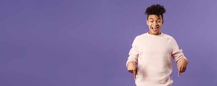 Joyful good-looking hipster guy with dreads rejoicing over good news, best deal ever in online store, favorite game finally in stock, pointing fingers down and rejoicing, purple background.