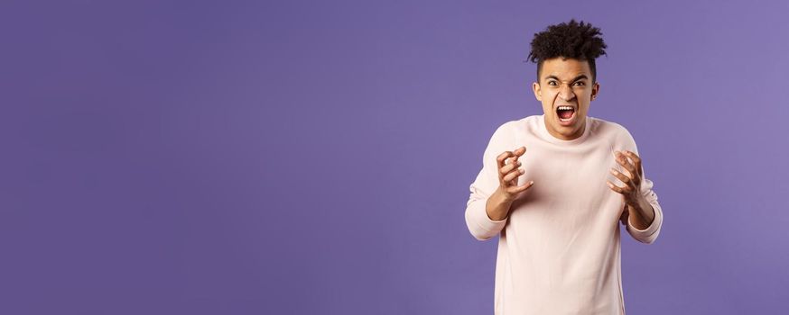 Portrait of hateful, outraged young hispanic man losing his temper, want to strangle or attack someone, cursing being aggressive, squeeze hands into fist from rage and hate, purple background.