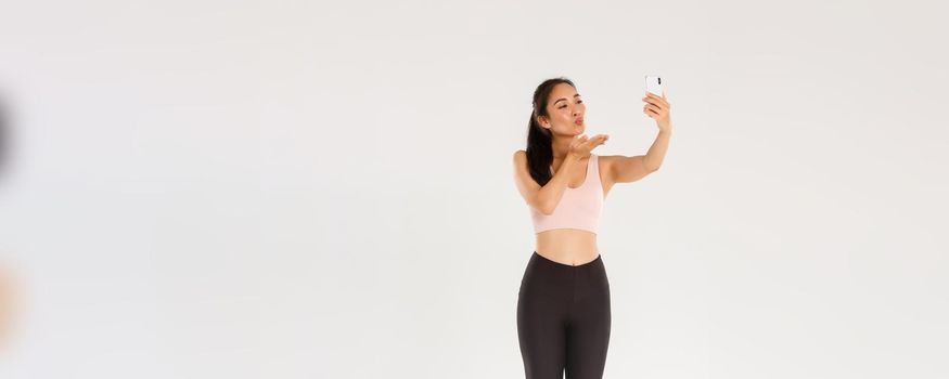 Full length of cute and silly female blogger in gym, wearing sportswear and taking selfie or live stream from gym workout session, sending air kiss at mobile phone screen, standing white background.