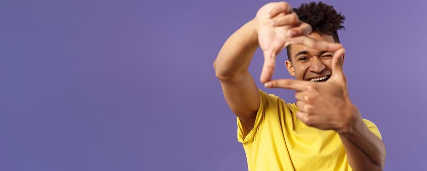 Close-up portrait of young creative photographer, man imaging, searching right angle, looking through photo frame, wink and smiling pleased, standing purple background found good shot.