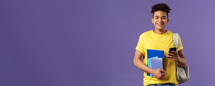 Back to school, university concept. Portrait of cheerful young handsome male student wearing backpack on shoulder, hold notebooks and studying books, texting friend mobile phone.