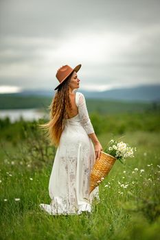 A middle-aged woman in a white dress and brown hat stands on a green field and holds a basket in her hands with a large bouquet of daisies. In the background there are mountains and a lake