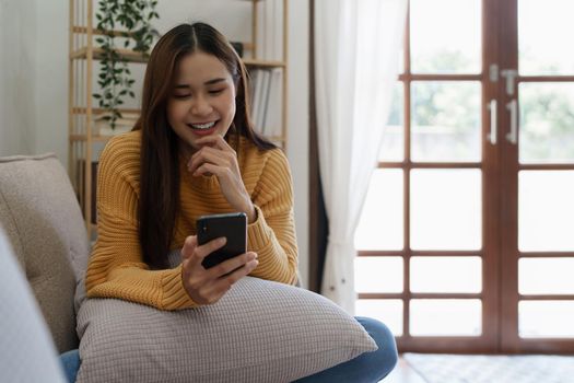 Cheerful asian young woman using mobile phone while sitting on a sofa at home.