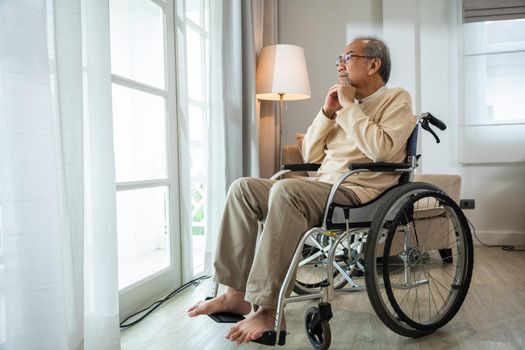 Asian senior man disabled sitting alone in wheelchair looking through window at hospital, lonely elder thoughtful sad old man look outside windows in bedroom at retirement home, feel depressed lonely