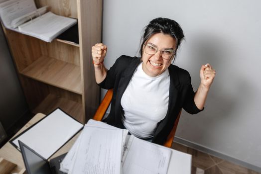 An angry office employee. The girl manager working with papers at her laptop is very angry and looks at the camera with anger. Stress at work, feeling unwell.