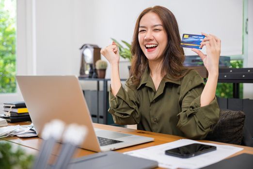 Happy Asian freelancer holding credit card she excited raised hands say yes! do winner gesture on desk home office, lifestyle business woman using laptop computer rejoicing to purchase shopping online