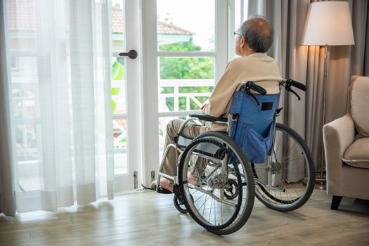 Asian senior man disabled sitting alone in wheelchair looking through window at hospital, lonely elder thoughtful sad old man look outside windows in bedroom at retirement home, feel depressed lonely