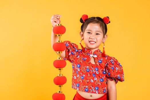 Chinese New Year. Happy Asian Chinese little girl smile wearing red cheongsam qipao holding silk lanterns on hand, Portrait children in traditional dress, studio short isolated on yellow background