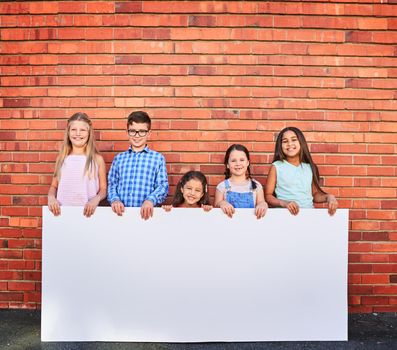 Well use our cuteness to help promote your message. Portrait of a group of young children holding a blank sign against a brick wall