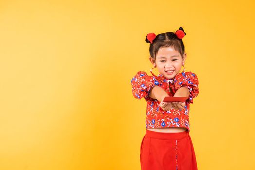 Chinese New Year. Happy Asian Chinese little girl smile wear red cheongsam handed angpao or red packet monetary gift, Portrait children in traditional dress, studio short isolated on yellow background